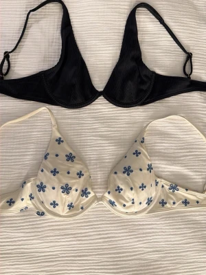 bikinitoppar från hollister  - Säljer två bikiniöverdelar från hollister i olika stilar. Båda har justerbara axelband, bygel och knäppe där bak. Den svarta är ribbad och utan vaddering, och den andra är vit med blå blommor och vaddering. Båda är flitigt använda men i bra skick, utan några defekter. Perfekta för dig som vill ha både basic och mönstrat i garderoben.