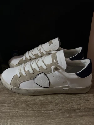 Philippe Model vita sneakers - Stilrena vita sneakers från Philippe Model med beige mockadetaljer och mörkblå häl. Klassisk låg modell med vita skosnören och logga på sidan. Yttermaterial i läder och mocka, perfekt för en clean och trendig look.