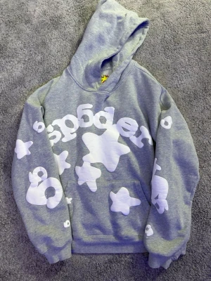 Grå hoodie från Sp5der med tryck - Grå hoodie från Sp5der med stora vita stjärn- och texttryck över hela tröjan. Hoodien har en klassisk huva och magficka framtill.