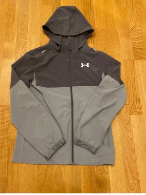 Grå vindjacka från Under Armour - Snygg tvåtonad grå vindjacka från Under Armour med huva och hel dragkedja framtill. Jackan har långa ärmar, elastiska muddar med logga och en vit logotyp på bröstet. Perfekt för blåsiga dagar och sportiga outfits.
