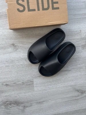 Svarta Adidas Yeezy Slides - Säljer ett par svarta Adidas Yeezy Slides med stilren och chunky design. Tofflorna är tillverkade i ett mjukt och flexibelt syntetmaterial som ger skön komfort. Perfekta för chill dagar hemma eller när du vill ha en clean look.