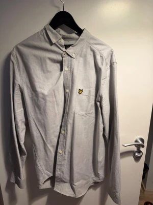 Ljusgrå skjorta från Lyle & Scott - Hej Säljer en Klassisk ljusgrå skjorta från Lyle & Scott med gul broderad logga på bröstfickan. Skjortan har knappar framtill, lång ärm och en stilren passform. Perfekt för dig som gillar enkel och snygg stil. Säljs Pga av För Liten som Ny Knappt Använd 💯 Inga Fläckar skador 