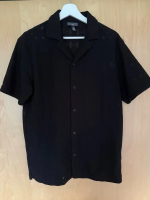 Svart kortärmad skjorta från H&M - Säljer en svart kortärmad skjorta från H&M med klassisk krage och knappar framtill. Skjortan har en luftig känsla och är perfekt för varmare dagar. Enkel och stilren design som passar till många olika outfits.