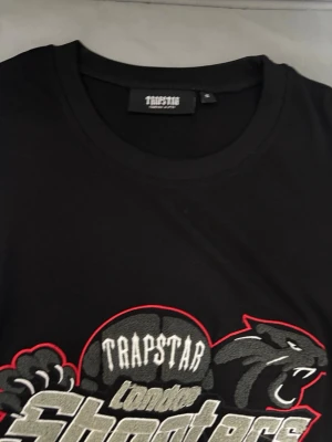 Svart Trapstar t-shirt med tryck - Snygg svart t-shirt från Trapstar med stort broderat tryck på bröstet där det står 'Trapstar London Shooters' och en panter. T-shirten har rund hals och korta ärmar. Perfekt för dig som vill ha en streetwear vibe.