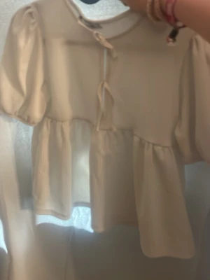 Krämvit blus med puffärm och knyt kostar 150 ny - Söt krämvit blus med puffiga kortärmar och volang nedtill. Blusen har två dekorativa knytband framtill och en lös, flowy passform. Materialet ser ut att vara ett mjukt tyg, troligen bomull eller viskos. Perfekt för en trendig och avslappnad look.
