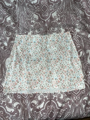 Vit blommig kortkjol - Söt vit kortkjol med små blommor i rosa, blått och grönt. Kjolen har en rak passform och är gjord i ett lätt och mjukt material, perfekt för varma dagar. Enkel att matcha med olika toppar för en fräsch look.