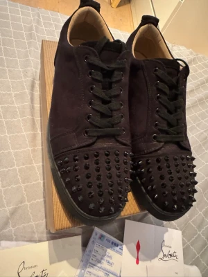Svarta spikes-sneakers Louboutin - Svarta sneakers från Christian Louboutin med mattsvarta nitar över tån och klassisk röd sula. Skorna har snörning och är tillverkade i mocka. Snygg och edgy design som sticker ut med sina detaljer.  Bara använda ett par gånger till speciella tillfällen men använder dom inte längre. Fick dom av en person som gav med kvitto 🧾och allt på bilden!!! Priset går att diskuteras 