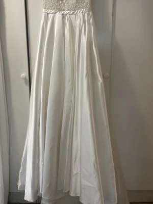 Vit bröllop långklänning med pärlor - Elegant vit långklänning med v-ringad överdel täckt av pärlor och smala axelband. Kjolen är i glansigt satinliknande material som faller mjukt. Perfekt för speciella tillfällen där du vill känna dig extra fin. Andra delar kommer med skriv om ni vill se dem! Storlek 38 men kan funka till lite större storlek för band grejen där bak