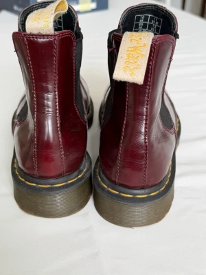Vinröda Chelsea boots Dr. Martens - Säljer ett par vinröda Chelsea boots från Dr. Martens i blankt läder. Klassisk modell med elastiska paneler på sidorna och den ikoniska gula sömmen runt sulan. Perfekta för dig som vill ha en edgy och tidlös look.