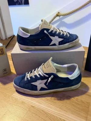 Golden goose superstars - Säljer ett par snygga blå Golden Goose superstars sneakers med vit stjärndetalj på sidan och vita skosnören. Skorna har beige plös och vit hälflik, samt en något smutsad sula för en cool, använd look. Ovandelen är i mocka och sulan är i gummi. Inga heelbites. Autentisering ingår samt kvitto. Nypris: 5500kr