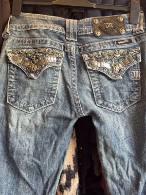 Blå bootcut Miss Me jeans med detaljer - Säljer ett par blå bootcut jeans från Miss Me med snygga broderade och glittriga detaljer på bakfickorna. Klassisk femficksmodell med låg midja och raka ben men även lite bootcut, ser väldigt raka ut på bilderna eftersom inte använda på ett tag. Jeansen har slitningar för en cool, avslappnad look och är tillverkade i denim. 