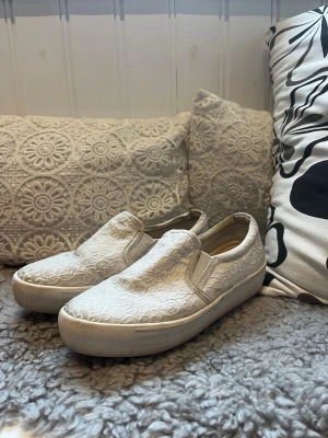 Vita slip-on sneakers med spetsmönster - Snygga vita slip-on sneakers med ett unikt spetsmönster över hela skon. Skorna har en tjock vit sula och elastiska paneler på sidorna för enkel på- och avtagning. Perfekta för dig som gillar en stilren men ändå detaljrik look.