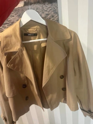 Kort beige trenchcoat från Gina Tricot - Snygg kort trenchcoat i beige från Gina Tricot. Jackan har dubbelknäppning med stora knappar, bred krage och justerbara band vid ärmsluten. Perfekt för dig som vill ha en klassisk men trendig look. Materialet känns mjukt och följsamt.