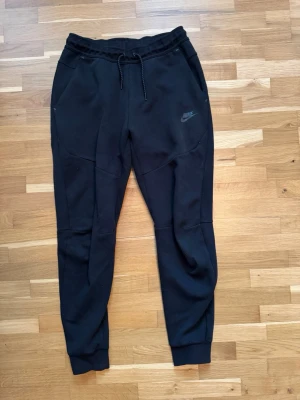 Svarta Techfleece byxor från Nike - Säljer ett par svarta mjukisbyxor från Nike med dragsko i midjan och ribbade muddar vid bensluten. Byxorna har två sidofickor fram och en ficka med dragkedja bak. Perfekta för chill eller träning.