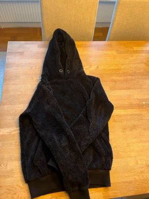 Svart fluffig hoodie med huva - Mysig svart hoodie i fluffigt teddy-material med stor huva och muddar vid ärmslut och nederkant. Perfekt för dig som gillar en avslappnad och trendig stil. Hoodien har en enkel design utan synligt tryck eller logga. Storlek S
