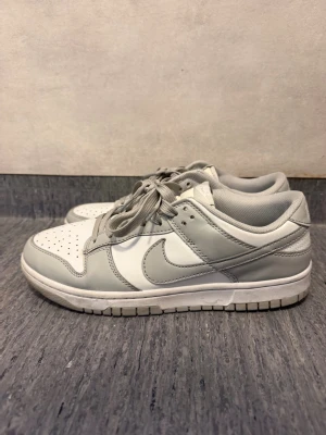 Gråvita Nike Dunk Low sneakers - Snygga Nike Dunk Low sneakers i vitt och ljusgrått läder. Klassisk siluett med perforerad tåbox, grå snörning och vit sula. Perfekta för dig som gillar stilrena och tidlösa sneakers med en sportig vibe.