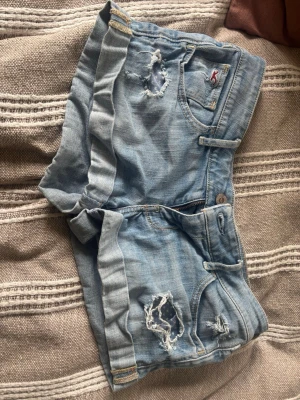 Ljusblå jeansshorts från Hollister - Säljer ett par ljusblå jeansshorts från Hollister med slitna detaljer och uppvikta benslut. Klassisk femficksmodell med broderad logga på fickan och bakfickor med dekorativa sömmar. Perfekta för sommaren!