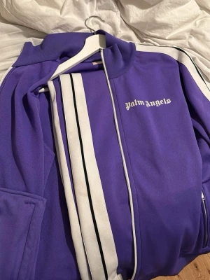 Lila tracksuit från Palm Angels - Säljer en lila tracksuit från Palm Angels. Både byxorna och tröjan säljs som ett komplett kit. Perfekt för en sportig och trendig look. Storlek L på tröjan och M på byxorna. 