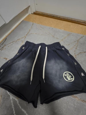 Svarta shorts med snörning från hellstar - Säljer ett par svarta shorts från hellstar med vit snörning i midjan och tryckt logga på ena benet. Shortsen har tryckknappar längs sidorna och är tillverkade i mjukt material, perfekta för chill dagar.
