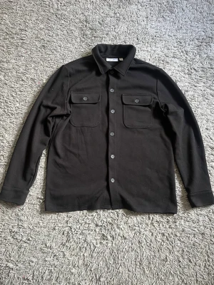 Svart overshirt från Only & Sons - Stilren svart overshirt från Only & Sons med klassisk krage och knäppning framtill. Två stora bröstfickor med lock och knapp ger en cool utility-vibe. Tillverkad i ett mjukt tyg som känns bekvämt och passar perfekt till lager-på-lager.