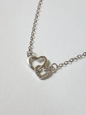 Silverhalsband med dubbla hjärtan - Snyggt halsband i silverfärg med två sammanlänkade hjärtan, där det ena hjärtat är dekorerat med små gnistrande stenar. Kedjan är tunn och smidig, perfekt för att ge en romantisk touch till din stil. Materialet rostfritt stål. 