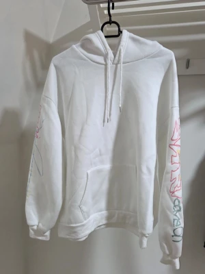 Vit hoodie med färgglad textprint - Säljer en vit hoodie med huva och känguruficka. Ärmarna har färgglada grafiska tryck och på ryggen står det 'LOVE' i stora bokstäver med flerfärgat tryck och textdetaljer. Hoodien har dragsko i huvan och ribbade muddar. Perfekt för dig som gillar streetwear och unika prints. Längd: ca 60 cm 