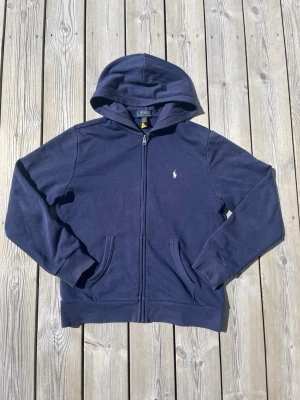 Marinblå hoodie från Polo Ralph Lauren - Klassisk marinblå hoodie från Polo Ralph Lauren med dragkedja framtill och vit broderad logga på bröstet. Hoodien har kängurufickor och ribbade muddar vid ärmar och nederkant. Tillverkad i mjuk bomullsblandning för skön komfort.