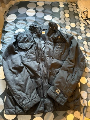 Svart parkajacka från Boss Orange - Snygg svart parkajacka från Boss Orange med flera praktiska fickor framtill och justerbara detaljer vid ärmslut och midja. Jackan har hög krage och dragkedja med knappslå. Perfekt för kyligare dagar och enkel att matcha.