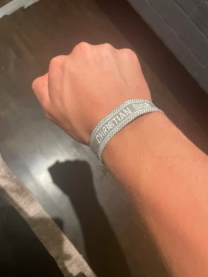 Grått Christian Dior tygarmband - Snyggt grått tygarmband från Christian Dior med broderad logga i vitt. Armbandet har fransade kanter och justerbar knytning för perfekt passform. Perfekt accessoar för dig som gillar exklusiva detaljer och vill lyfta din outfit med en ikonisk touch.