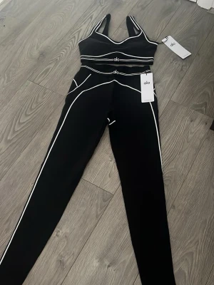Svart träningsset från Alo och Lululemon - Två olika träningsset: ett svart set från Alo med vita kontrastsömmar, sport-bh och tights med hög midja, samt ett svart set från Lululemon med jacka med gulddragkedja och matchande tights. Båda seten är i stretchigt funktionsmaterial, perfekta för gym eller yoga.