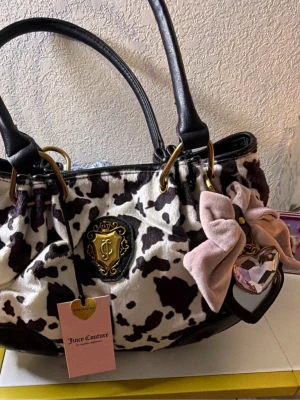 Juicy Couture handväska med ko-mönster - Juicy Couture handväska i plysch med svartvitt ko-mönster, svarta handtag i konstläder och guldfärgade detaljer. Väskan har en dekorativ rosett och hjärtberlock samt ett emblem med JC-logga framtill. Perfekt statement piece för dig som gillar unika accessoarer.