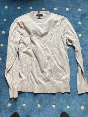 Beige stickad tröja Tommy Hilfiger - Snygg beige stickad tröja från Tommy Hilfiger med rund halsringning och diskret logga på bröstet. Tröjan har långa ärmar, ribbade muddar och klassisk passform. Perfekt till jeans eller chinos för en clean look.