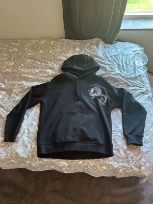 Svart hoodie från HUGO - Snygg svart hoodie från HUGO med stor huva och känguruficka. På bröstet finns ett tryck med två vita rosor och HUGO-loggan i svart. Hoodien har ribbade muddar och är gjord i mjukt bomullsmaterial. Perfekt för en chill och stilren look.