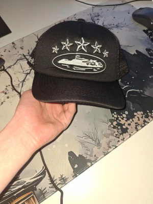 Svart corteiz keps - Säljer en svart keps med mesh på sidorna och broderade vita stjärnor samt ett ovalt motiv framtill. Kepsen har böjd skärm och justerbar baksida. Perfekt för dig som gillar streetwear och vill sticka ut med coola detaljer.