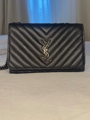 Svart YSL quiltad handväska - Säljer en svart quiltad handväska från Saint Laurent med ikoniskt YSL-emblem i silver på framsidan. Väskan har en stilren, rektangulär form och kedjerem i metall. Tillverkad i slätt läder med snyggt mönster i chevron-stil. Perfekt för dig som gillar lyxiga accessoarer.