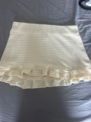 Gulvit randig kjol med volang från Nelly - Supersöt kort kjol från Nelly i gulvitt rutigt mönster. Kjolen har volangdetaljer längst ner som ger en extra feminin touch. Kjolen är i stretchigt material med resorband i midjan och säljs inte längre. Aldrig använd men prislappen är borttagen vilket gör den ny utan prislapp