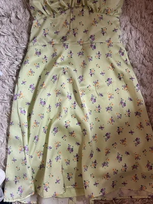Ljusgrön blommig klänning med snörning - Söt ljusgrön klänning med små blommor i lila och gult. Klänningen har snörning och dragkedja, vilket ger en trendig look. Perfekt för varma dagar och sommarens alla tillfällen. Materialet känns lätt och luftigt.