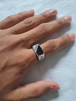 Silverfärgad ring med svart sten - Snygg silverfärgad ring med en stor, rektangulär svart sten i mitten. Ringen har detaljer i form av små triangulära mönster med glittrande stenar på ena sidan. Modern och stilren design som sticker ut.