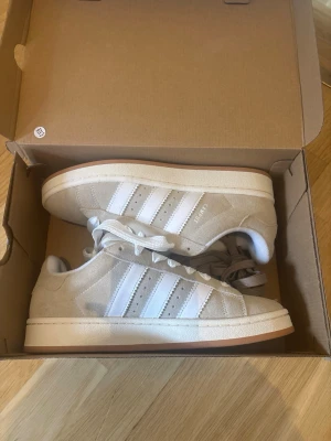 Helt nya Adidas Campus 00s Beige/grå  - Säljer ett par helt nya och oanvända Adidas Campus 00s i snygg beige/vit färg. Skorna är endast uppackade och provade inomhus, aldrig använda utomhus. Normalt säljs skorna för cirka 1500kr  ✅ Helt nya ✅ Originalkartong medföljer ✅ Extra bruna skosnören medföljer ✅ Trendig och lättmatchad färg  Storlek: 40 2/3 Skick: Helt nya, oanvända Färg: Beige/grå Modell: Adidas Campus 00s Helt nya trendiga sneakers med originalkartong och extra bruna skosnören. Oanvända.