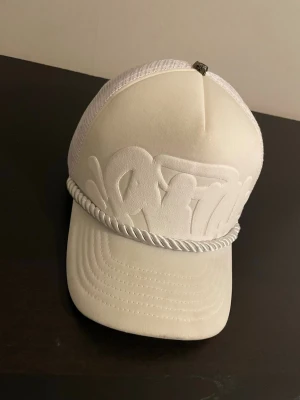 Vit keps från Syna World - Säljer en vit keps från Syna World med broderad logga framtill och coolt repdetalj ovanför skärmen. Kepsen har mesh på sidorna och justerbar baksida för perfekt passform. Materialet är en mix av bomull och polyester, vilket gör den både bekväm och luftig.