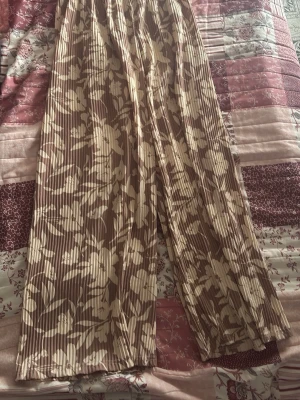 Plisserade blommiga leggings beige/brun - Säljer ett par plisserade leggings med beige och brunt blommönster. De har bred resår i midjan för skön passform och är tillverkade i ett lätt, stretchigt material. Perfekta för dig som gillar en avslappnad men trendig stil.