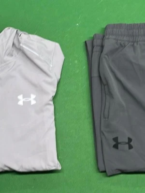 Under Armour träningsset grå - Snyggt träningsset från Under Armour med en ljusgrå ziptröja och mörkgrå byxor. Båda plaggen har Under Armour-loggan framtill. Tröjan har dragkedja vid halsen och byxorna har resår i midjan. Perfekt för gymmet eller löprundan.