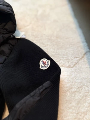 Svart Moncler jacka  - Säljer en svart pufferjacka från Moncler med quiltad kropp och stickade ärmar. Jackan har en stor huva, dragkedja framtill och Moncler-logga på ena ärmen. Perfekt för kalla dagar och riktigt snygg streetstil.