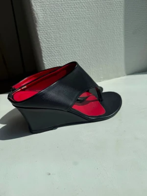 Svarta kilklackade sandaler - Snygga svarta sandaler med kilklack och öppen tå. Insidan är röd för en cool kontrast. Sandalerna har breda remmar i konstläder och dragkedja bak för enkel påtagning. Perfekta för dig som vill ha både höjd och stil.