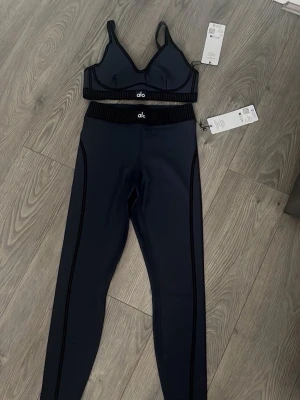 Mörkblått träningsset från Alo - Snyggt mörkblått träningsset från Alo med sport-bh och höga tights. Setet har svarta detaljer och bred resår i midjan och under bysten. Perfekt för gymmet eller yoga. Materialet är stretchigt och följsamt för maximal komfort. Är tillgänglig i alla storlekar och det kommer som ett sett !! 