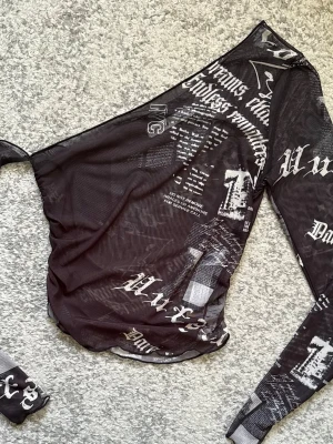 Svart mesh topp med tryck och lång ärm - Säljer en svart mesh topp med vita tryck i olika typsnitt och mönster. Toppen har lång ärm och är gjord i ett tunt, transparent material. Perfekt för att skapa en edgy och trendig look.