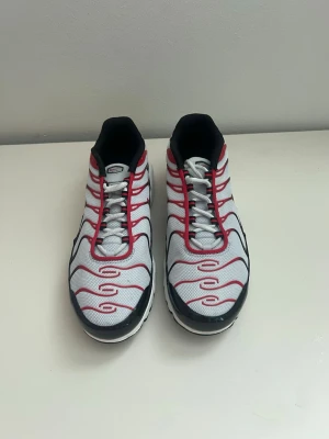 Nike air max Plus vita/röda sneakers - Nike Air Max Plus sneakers i vitt mesh med röda och svarta detaljer. Skorna har den ikoniska Tuned Air-sulan, röda vågformade linjer och svart yttersula. Swoosh-logga på sidan och klassisk snörning. Perfekta för dig som gillar streetwear och sportig stil.
