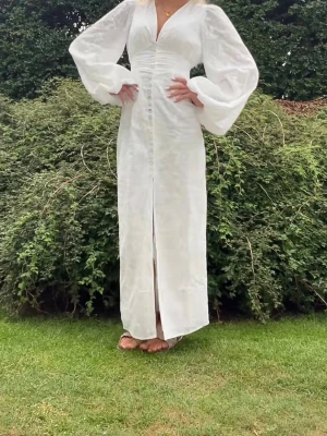 Vit långklänning med puffärm Adoore  - Elegant vit långklänning med v-ringning, puffiga långärmar och knappar framtill. Klänningen har en slits framtill och markerad midja som ger en feminin siluett. Perfekt för sommarens alla tillfällen.