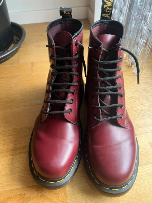 Dr. Martens  - Snygga vinröda boots i läder med klassisk snörning och robust sula med gul söm. Högt skaft och svarta detaljer, inklusive svarta skosnören och dragflik baktill. Perfekta för att lyfta din stil med en edgy touch.