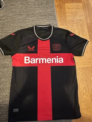Bayer Leverkusen matchtröja svart/röd - Snygg Bayer Leverkusen matchtröja från Castore i svart med röd korsdesign och vita detaljer. Tröjan har klubbmärke och sponsortryck på bröstet samt rund halsringning. Perfekt för fotbollsfans som vill sticka ut på läktaren eller planen.